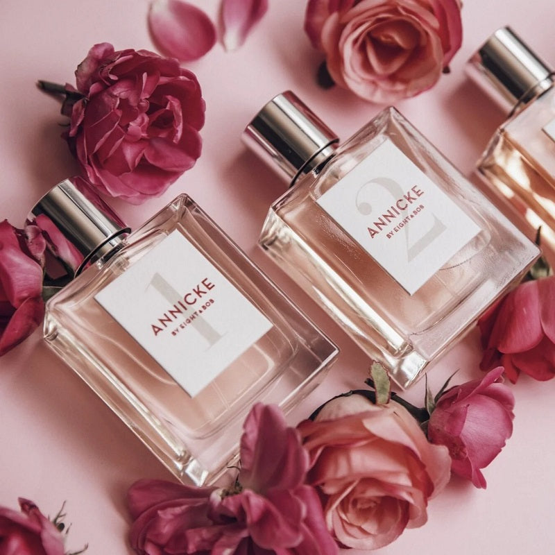 Two Annicke perfume bottles surrounded by pink roses on a pink background