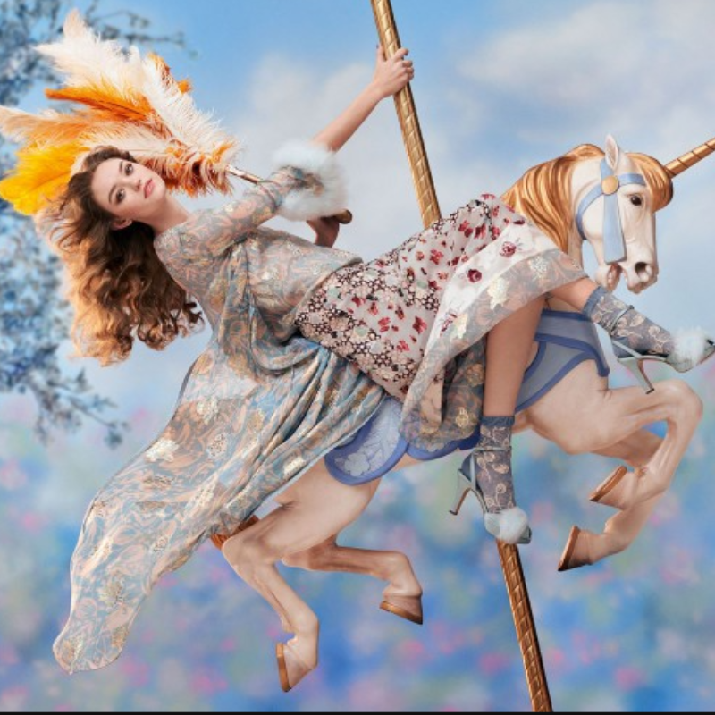 Woman in a floral dress riding a carousel unicorn against a blue sky.