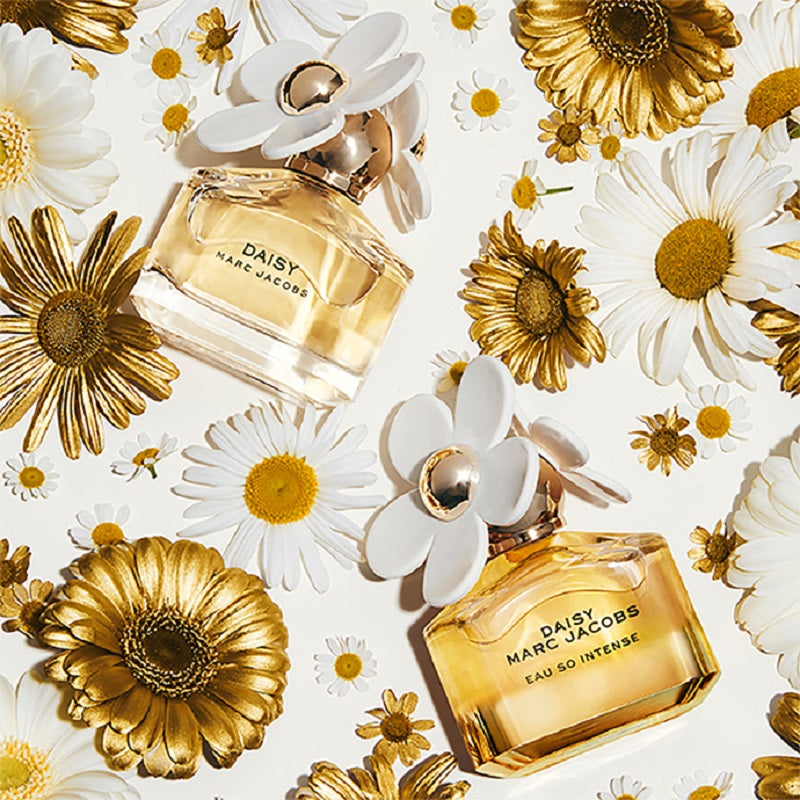 Two bottles of Marc Jacobs Daisy perfume surrounded by white and gold flowers on a light background