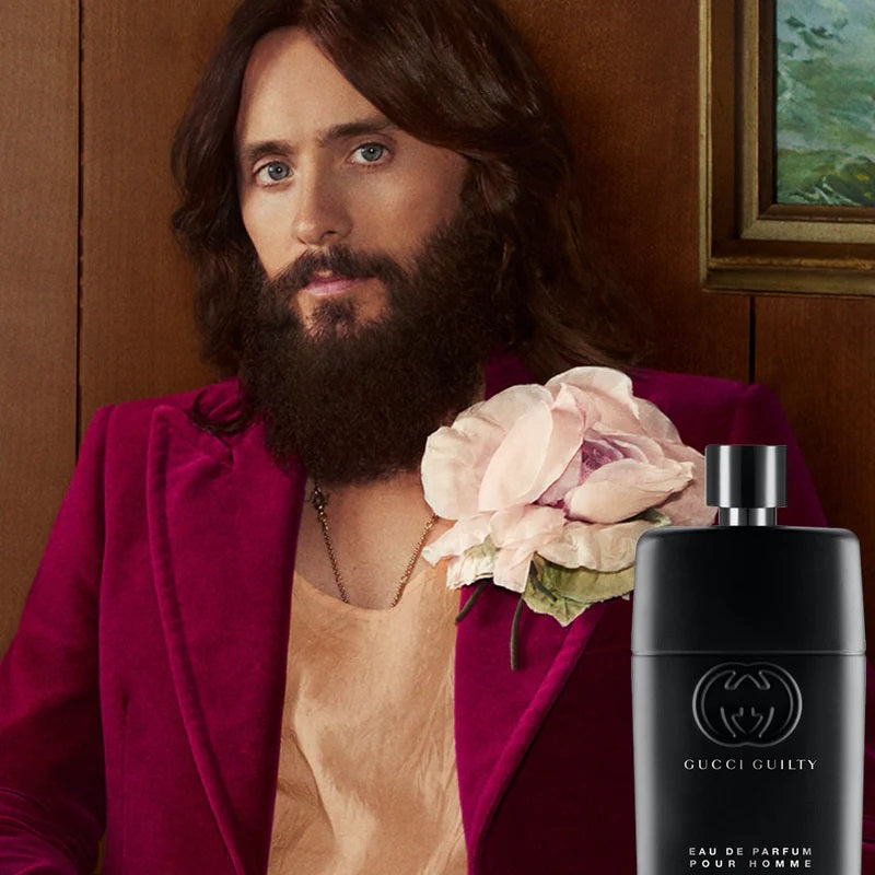 Man with a beard wearing a red jacket with a flower, next to a Gucci perfume bottle.
