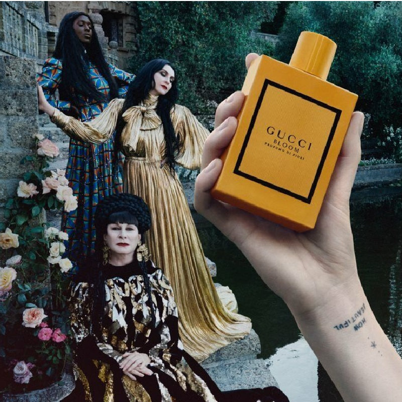 Hand holding a Gucci Bloom perfume bottle in front of a collage with women in vintage attire and flowers.