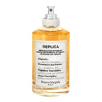 A bottle of 'By the Fireplace' Eau de Toilette by Maison Margiela, with a clear, amber-colored liquid visible and a label that includes the product name and a brief description.