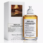 A product image featuring a 100ml bottle of 'By the Fireplace' Eau De Toilette by Maison Margiela, with a white box packaging displaying a fireplace image.