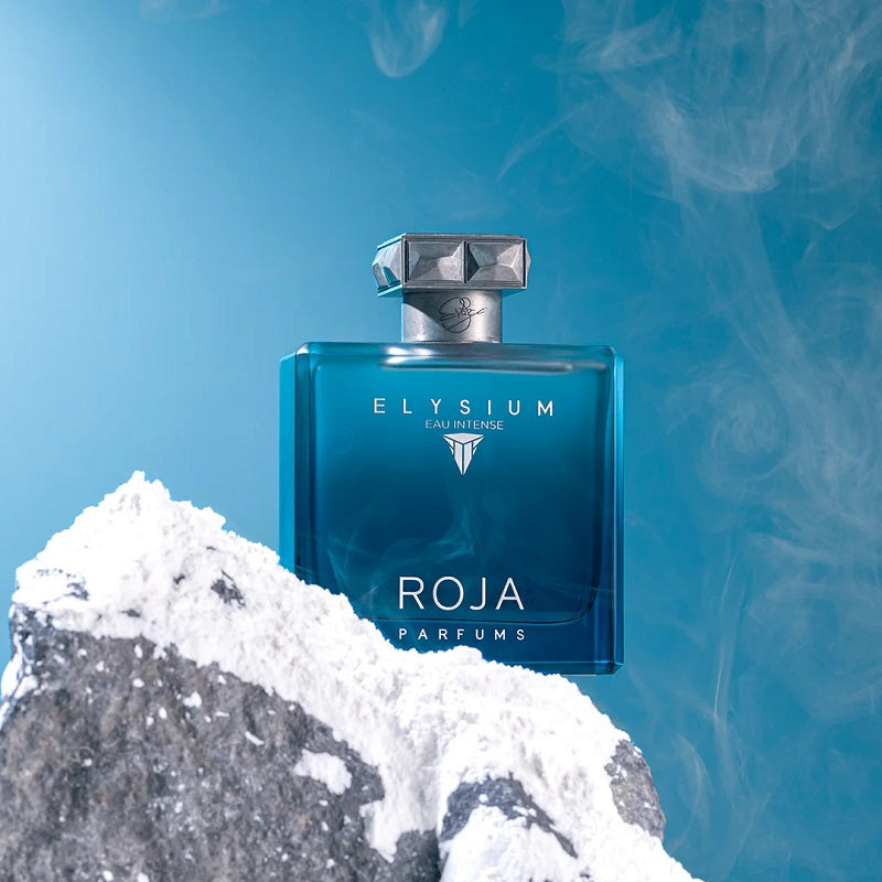 Blue ROJA ELYSIUM perfume bottle on a rocky surface with a blue background