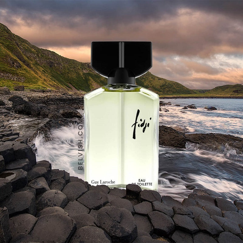Perfume bottle labeled 'Fiji' on a rocky beach with ocean and mountains in the background