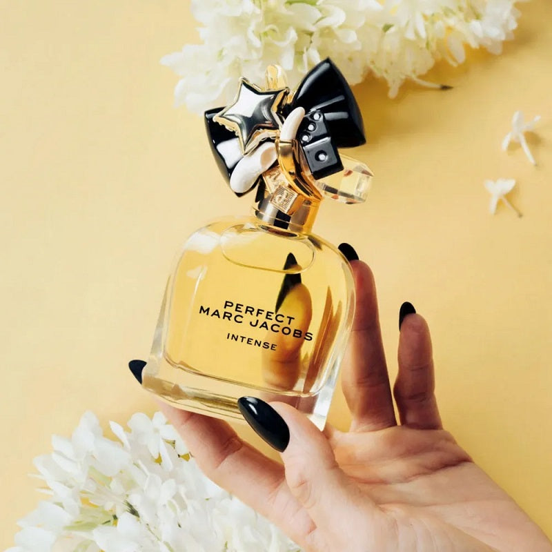 Hand holding a gold Marc Jacobs Perfect Intense perfume bottle against a yellow background with white flowers.