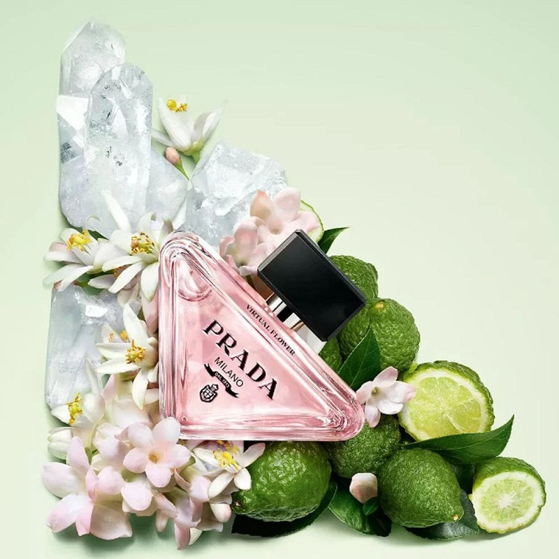 Prada Sempere Florence perfume bottle in a triangular pink glass design, surrounded by white flowers, green limes, and ice crystals on a pale mint green background.