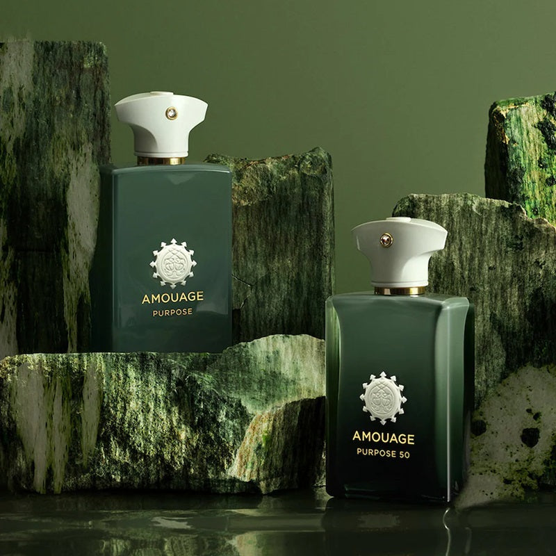 Two Amouage Purpose perfume bottles in dark green glass with white caps, displayed on textured green stone blocks against a matching olive green background.