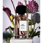 Gris Charel Eau de Parfum bottle with a black cap and white label, styled in a floral arrangement with purple flowers and green leaves on a light background.