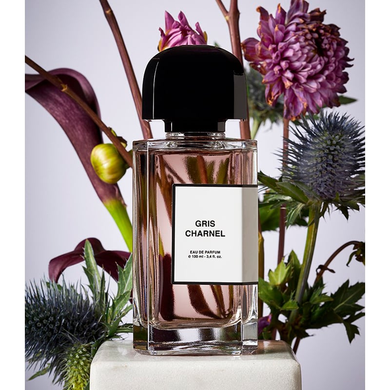 Gris Charel Eau de Parfum bottle with a black cap and white label, styled in a floral arrangement with purple flowers and green leaves on a light background.