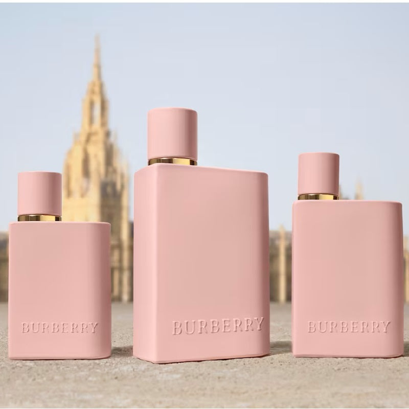 Three pink Burberry perfume bottles on a beach with a blurred architectural structure in the background.