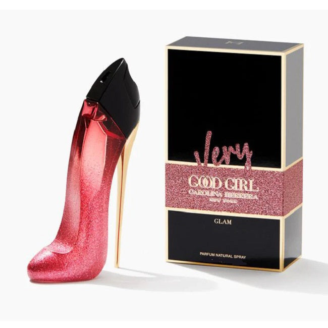 A product image featuring a bottle of 'Very Good Girl Glam' Eau De Parfum by Carolina Herrera, alongside its packaging. The bottle is designed to resemble a high-heeled shoe and is glittery pink, with the packaging box is black with gold and pink text and design elements.