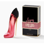 A product image featuring a bottle of 'Very Good Girl Glam' Eau De Parfum by Carolina Herrera, alongside its packaging. The bottle is designed to resemble a high-heeled shoe and is glittery pink, with the packaging box is black with gold and pink text and design elements.