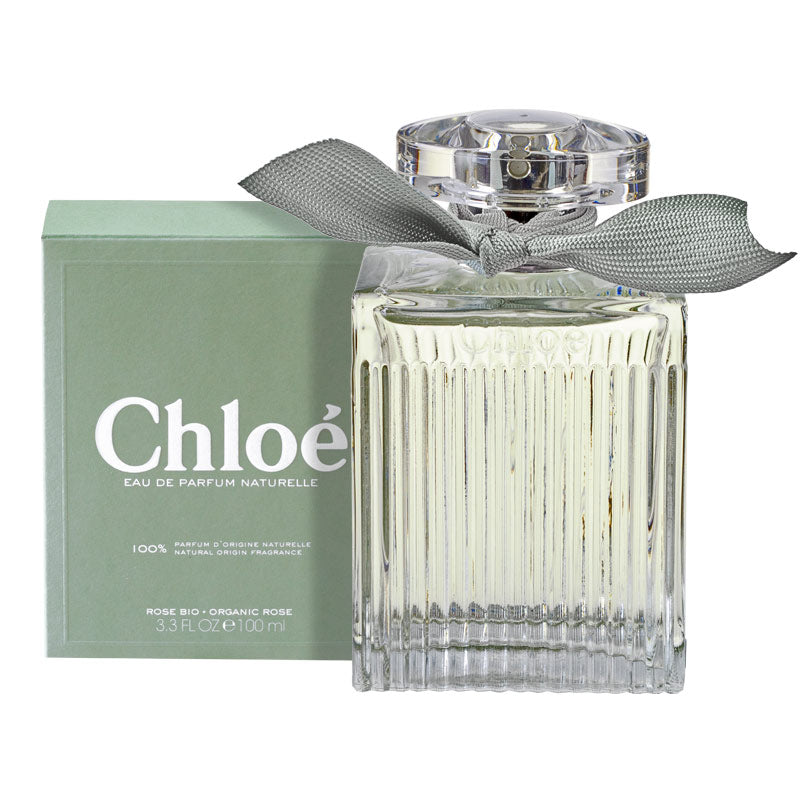 A bottle of Chloé Eau De Parfum Naturelle with a clear, beaded design and a silver cap, placed in front of its green box with a decorative bow.