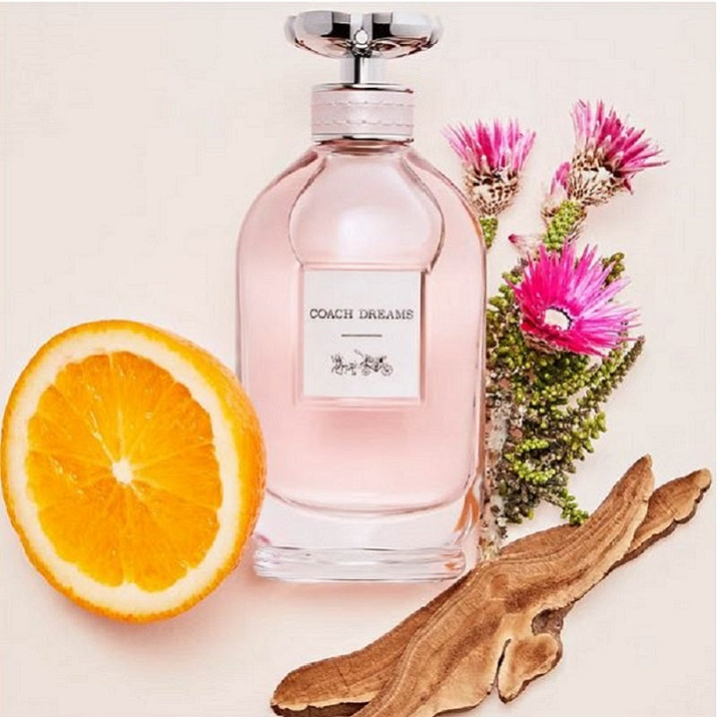 Coach Dreams perfume bottle with an orange, wooden piece, and flowers on a light background
