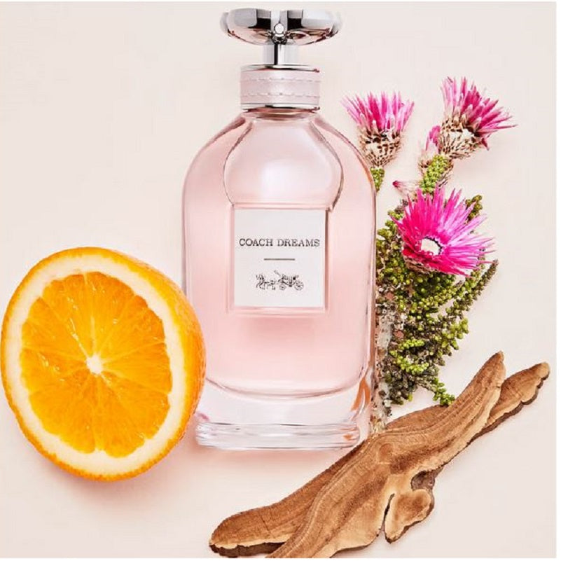 Coach Dreams perfume bottle with an orange, wood piece, and flowers on a light background