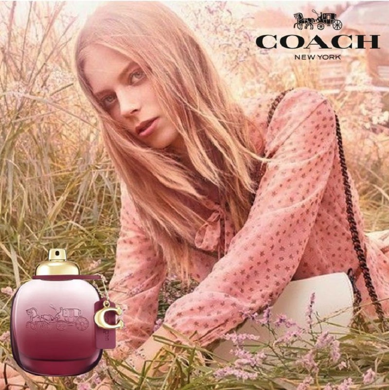 Woman in a pink dress sitting in a field with a Coach perfume bottle.