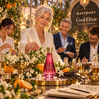 A woman in a cream blazer stands behind a decorated table displaying a Davidoff Cool Elixir bottle, surrounded by oranges, white flowers, vanilla beans, and other fragrance bottles in a warm, elegant setting. Two men and another woman observe in the background under hanging lights.
