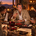 A man in a beige jacket sits at a table in a warm, rustic setting, holding a small red crystal. In the foreground, a dark red glass bottle labeled 'DAVIDOFF COOL ELIXIR ALFONSO MINERAL' sits on the table, surrounded by mineral samples, brass tools, and another person holding a crystal cluster.
