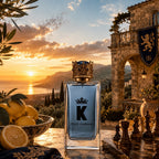 A Dolce & Gabbana King perfume bottle with a gold crown cap stands on a stone terrace at sunset, with lemons in a bowl, chess pieces, and a stone archway in the background.