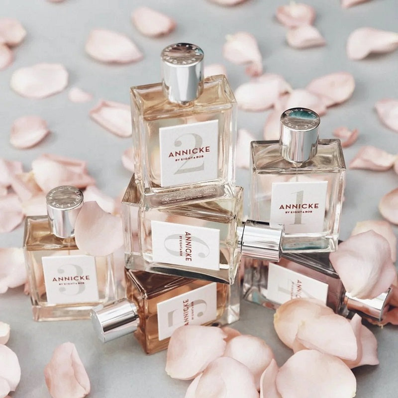 Packs of Annicke perfume bottles surrounded by pink petals on a light gray background