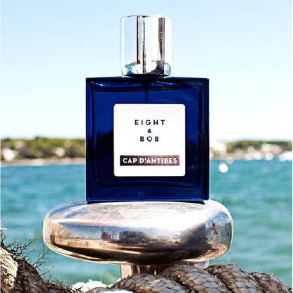 Blue perfume bottle labeled 'Eight & Bob Cap d'Antibes' on a rocky surface with water and sky in the background