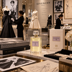 A Givenchy III perfume bottle with a gold cap and pale yellow liquid, displayed on a marble counter in a luxury Givenchy Paris boutique alongside branded books, sketches, and mannequins.