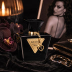 GUESS Seductive Noir perfume bottle in glossy black glass with a gold triangular logo plate and gold chain detail, styled on dark fabric with flowers, a candle, and a blurred woman in the background.
