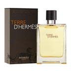 A bottle of Terre d'Hermes Eau de Toilette by Hermès, with a box in the background. The bottle is transparent, showcasing the yellowish liquid inside, with a black cap.