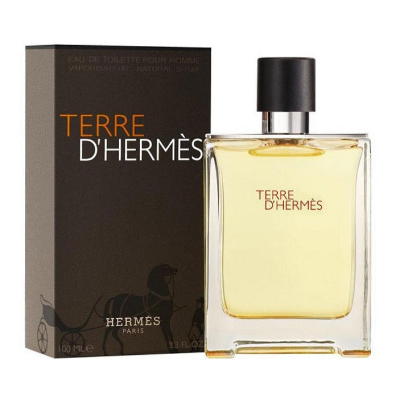 A bottle of Terre d'Hermes Eau de Toilette by Hermès, with a box in the background. The bottle is transparent, showcasing the yellowish liquid inside, with a black cap.