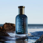 A bottle of Boss Bottled Infinite Eau de Parfum by Hugo Boss, positioned on a wet surface with a natural background featuring rocks and sky.