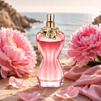 A pink ribbed perfume bottle with a gold cap and floral ornament, set on a beach with pink flowers and petals around it.