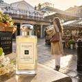 Jo Malone London 'Melrose Blossom & Honey' perfume bottle displayed on a stone ledge in Covent Garden Market, with a woman walking in the background.