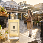 Jo Malone London 'Melrose Blossom & Honey' perfume bottle displayed on a stone ledge in Covent Garden Market, with a woman walking in the background.