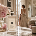 Jo Malone London Peony & Blush Suede Cologne perfume bottle in the foreground, styled on a marble table next to pink peony flowers, with a woman in a blush gown standing in a luxurious room in the background.
