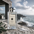 Jo Malone London Wood Sage & Sea Salt Cologne in a clear glass bottle with a silver cap, set on a rocky seaside cliffside with ocean waves and a modern building in the background.