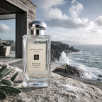 Jo Malone London Wood Sage & Sea Salt Cologne in a clear glass bottle with a silver cap, set on a rocky seaside cliffside with ocean waves and a modern building in the background.