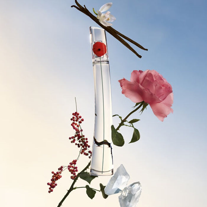 Kenzo Flower Eau de Parfum bottle surrounded by a pink rose, red berries, white blossoms and dark vanilla pods against a softly lit blue and cream background. 