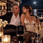 A man and a woman sit closely together on a yacht at night, with a black perfume bottle and box labeled Kilian on the table between them, alongside lit candles, flowers, and champagne glasses.