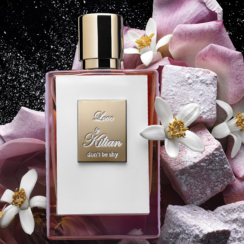 A rectangular perfume bottle labeled 'Love by Kilian don't be shy' with a metallic gold cap, surrounded by pink roses, white blossoms, and pink stone cubes on a dark sparkling background.