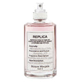 A clear spray bottle labeled 'REPLICA' by Maison Margiela, with text describing the fragrance as a reproduction of familiar scents and moments. The bottle is filled with a pinkish liquid.