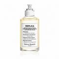 A clear glass perfume bottle with a white spray top and a white front label reading 'REPLICA' and 'Maison Margiela PARIS'. The bottle contains a pale yellow liquid and is photographed on a clean white background with a faint reflection below.