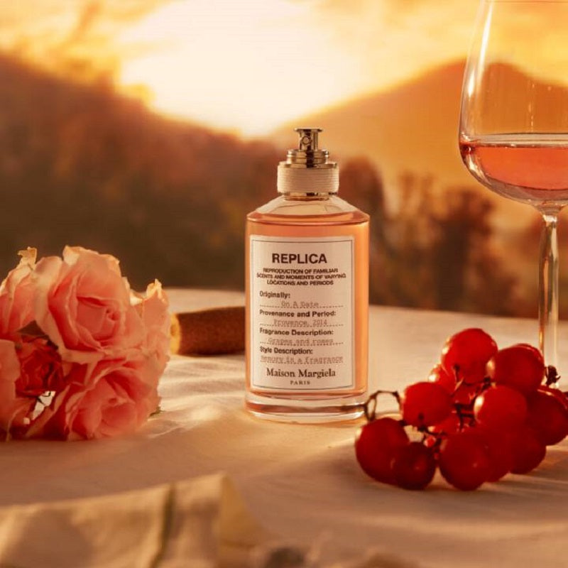 Perfume bottle labeled 'REPLICA' with a glass of wine, roses, and grapes on a sunset background.