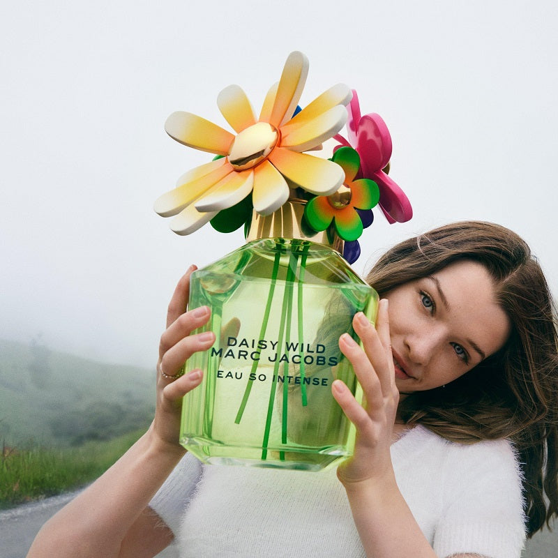 Person holding a large green perfume bottle with colorful flowers on top, set against a scenic background.
