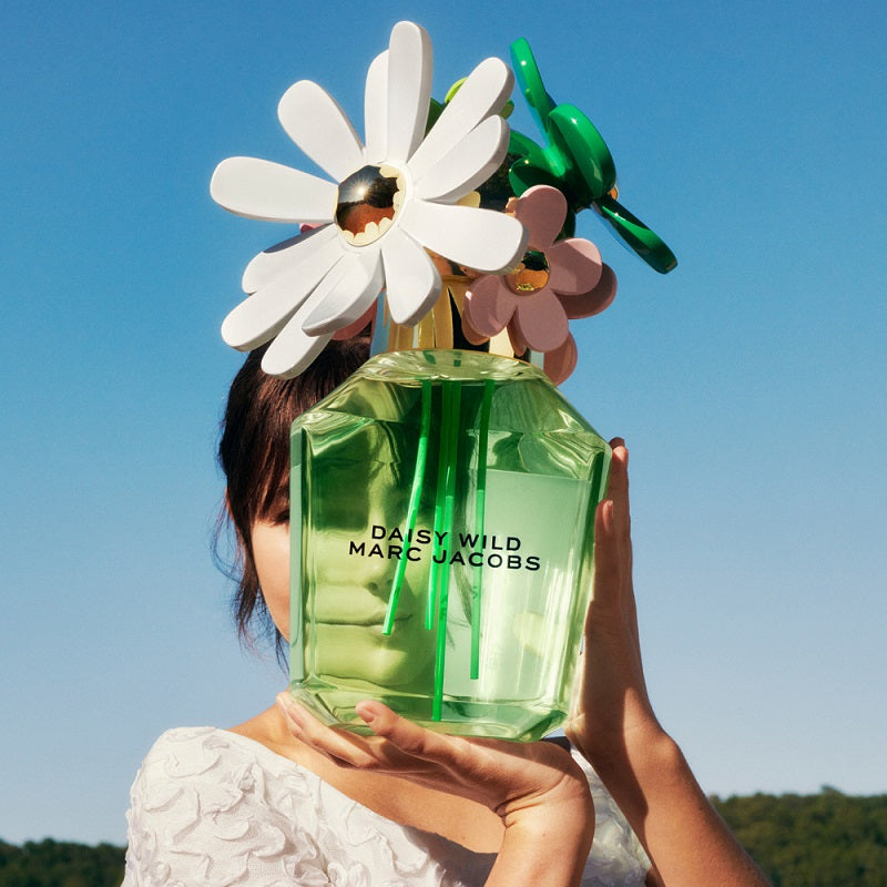 Person holding a green Marc Jacobs Daisy Wild perfume bottle with flowers against a blue sky.