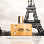 A perfume bottle labeled 'Eau de MEMO PARIS' with a gold cap and a gold label decorated with black-and-white birds, set against a blurred Eiffel Tower background.
