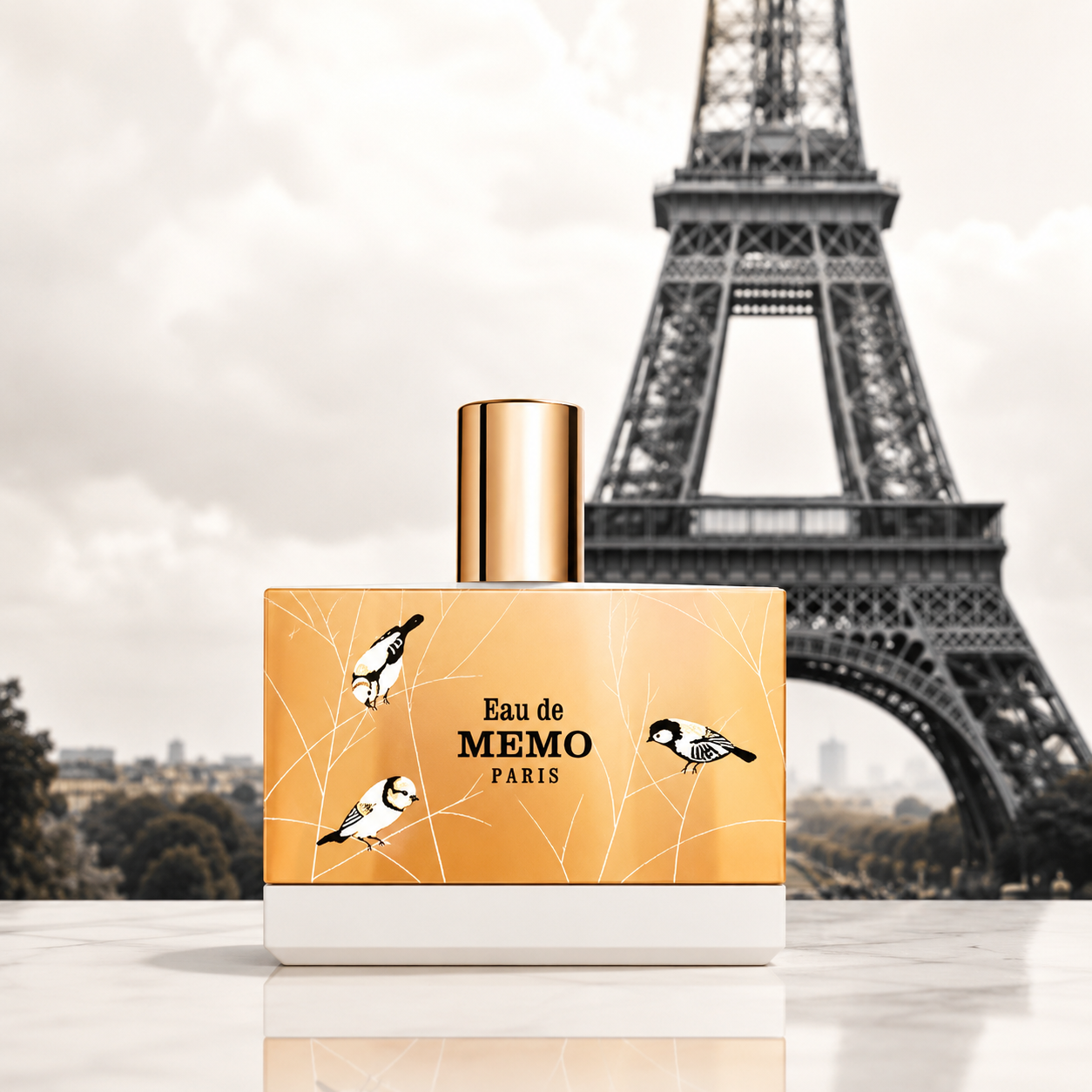 A perfume bottle labeled 'Eau de MEMO PARIS' with a gold cap and a gold label decorated with black-and-white birds, set against a blurred Eiffel Tower background.