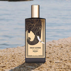 Perfume bottle labeled 'Indian Leather' by Memo on a sandy beach with water in the background