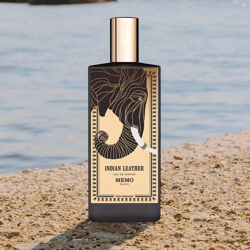 Perfume bottle labeled 'Indian Leather' by Memo on a sandy beach with water in the background