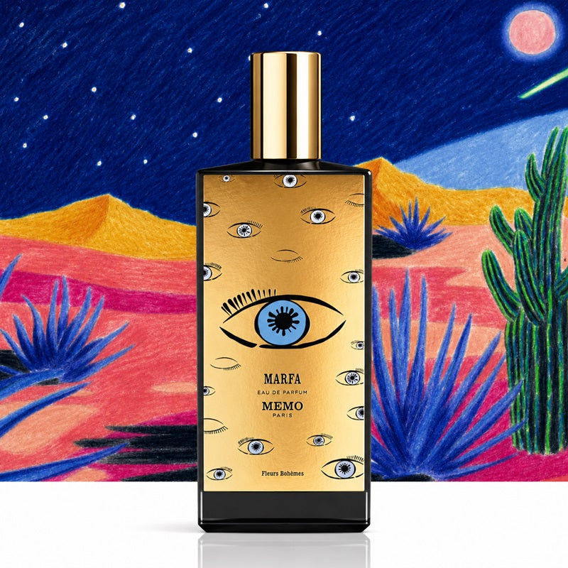 A rectangular perfume bottle with a gold front panel and black base stands centered against a colorful illustrated desert night scene. The bottle is labeled MARFA MEMO PARIS, with repeated eye motifs on the label and a shiny gold cap.